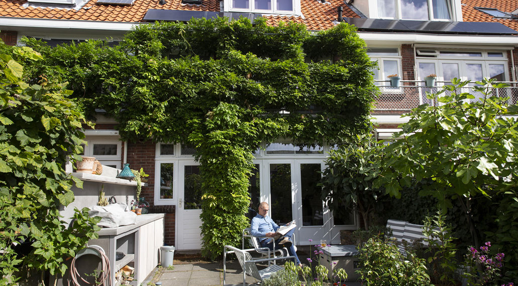 Een man leest in een tuinstoel, in zijn tuin staan bloeiende platen en de gevel is bijna helemaal begroeid om hittestress tegen te gaan.